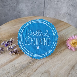 Könnte beinhalten: Ein runder, blau bestickter Aufnäher mit den Worten "Endlich SCHULKIND" in weißer Schrift und einem kleinen weißen Herz. Der Aufnäher hat einen gewellten Rand und liegt auf einer Holzoberfläche mit getrockneten Blumen.