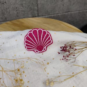 Könnte beinhalten: Ein gestickter rosa Glitzer-Muschel-Patch mit weißem Umriss. Der Patch befindet sich auf einem weißen Stoffhintergrund mit goldenem Glitzer.