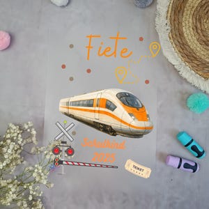 Könnte beinhalten: Eine transparente Folie mit einer Zugillustration, dem Namen "Fiete" und dem Text "Schulkind 2025". Der Zug ist weiß und orange, mit einem Ticket und einem Bahnübergang.