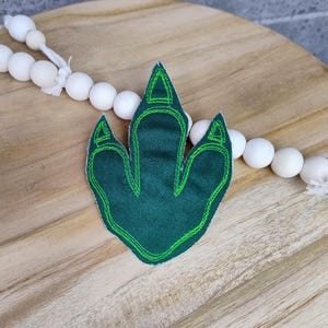 Könnte beinhalten: Grüner Dinosaurier-Fußabdruck-Patch mit einem lindgrünen Umriss. Der Patch ist aus Filz und perfekt, um jedem Projekt eine lustige Note zu verleihen.