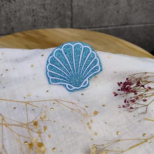 Könnte beinhalten: Ein hellblauer Glitzer-Muschel-Patch mit weißem Umriss. Der Patch befindet sich auf einem weißen Stoffhintergrund mit goldenen Glitzerpunkten.