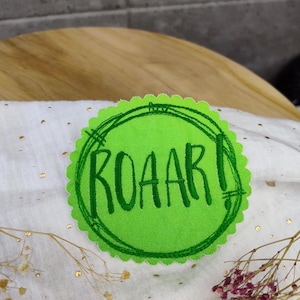 Könnte beinhalten: Grüner Filz-Kreis-Patch mit weißem Rand und dem in Grün gestickten Wort "ROAR!"