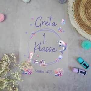 Könnte beinhalten: Eine transparente Folie mit dem Namen "Greta" und dem Text "1. Klasse" in Lila, umgeben von einem Meeresdesign mit Delfin, Seepferdchen und Seesternen. Der Text "Schulkind 2026" ist ebenfalls vorhanden.