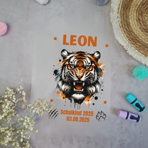 Könnte beinhalten: Eine transparente Tüte mit einer Illustration eines Tigers in Orange, Schwarz und Weiß. Der Name "LEON" ist über dem Tiger aufgedruckt, darunter steht "Schulkind 2025 03.08.2025". Die Tüte hat einen Pfotenabdruck und Kratzspuren.