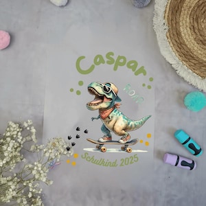 Könnte beinhalten: Hellgraue Grafik mit einem Cartoon-Dinosaurier auf einem Skateboard, der eine Sonnenbrille und ein Halstuch trägt. Der Text "Caspar" und "Schulkind 2025" sind ebenfalls vorhanden. Der Dinosaurier ist türkis und beige.