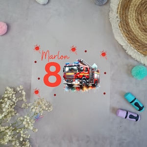 Könnte beinhalten: Ein rotes Feuerwehrauto mit einem weißen und blauen Aquarellhintergrund. Die Zahl 8 ist in Rot mit dem Namen Marlon darüber. Es gibt rote Farbspritzer auf einem grauen Hintergrund.