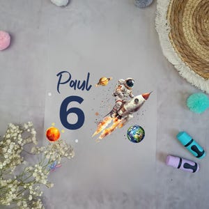 Könnte beinhalten: Eine transparente Folie mit Weltraum-Motiv. Sie zeigt einen Astronauten auf einer Rakete, Planeten und den Namen "Paul" über der Zahl "6". Das Design umfasst orange, blaue und weiße Elemente auf grauem Hintergrund.