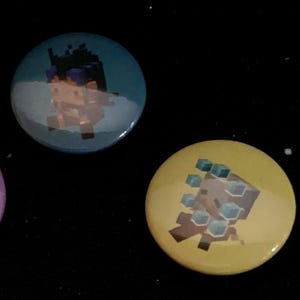 Op de afbeelding: Drie ronde knopen met pixelachtige karakterontwerpen. E&eacute;n knoop is paars met een personage dat een gele kubus vasthoudt, een andere is blauwgroen met een personage met donker haar, en de laatste is geel met blauwe accenten.