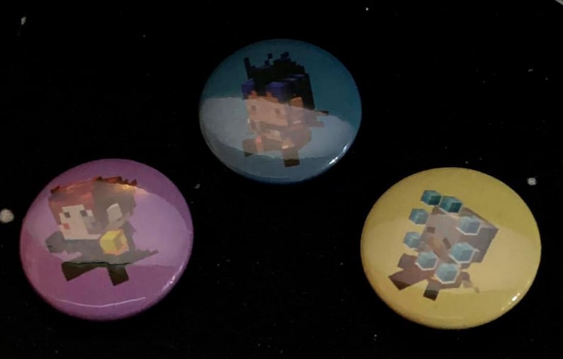 Op de afbeelding: Drie ronde knopen met pixelachtige karakterontwerpen. E&eacute;n knoop is paars met een personage dat een gele kubus vasthoudt, een andere is blauwgroen met een personage met donker haar, en de laatste is geel met blauwe accenten.