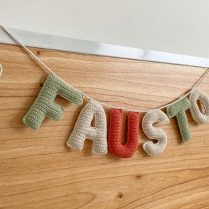 May include: A crocheted banner with the name "FAUSTO" spelled out in green, beige, and orange letters. The banner is strung with white yarn and is hanging from a wooden surface.