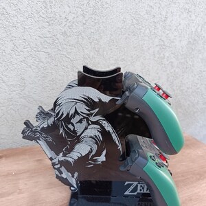 May include: Black acrylic video game controller stand featuring a graphic of a character from The Legend of Zelda. The stand holds two controllers with green and black colour scheme. The text "ZELDA" is visible on the base.