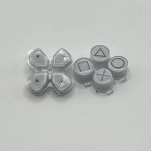 May include: A set of gray replacement buttons for a gaming controller. The set includes a directional pad and four individual buttons with symbols: a triangle, a square, a circle, and a cross. The buttons are made of a translucent plastic.