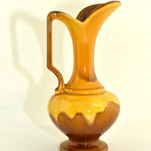 May include: A ceramic pitcher with a handle, featuring a two-tone glaze. The upper portion is a warm, golden-yellow, transitioning to a rich, dark brown at the base. The pitcher has a flared spout and a pedestal base.