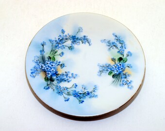 Puede incluir: Un platillo de porcelana blanca con un diseño floral azul y verde. Las flores son nomeolvides y están dispuestas en un patrón circular alrededor del borde del platillo.