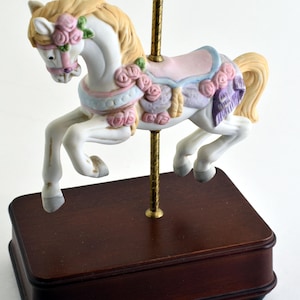 Puede incluir: Figura de caballo de carrusel con cuerpo blanco, adornado con rosas rosas y una silla de montar rosa claro. El caballo tiene crin y cola rubias, montado sobre una base de madera marrón oscuro. Un poste dorado sostiene el caballo.