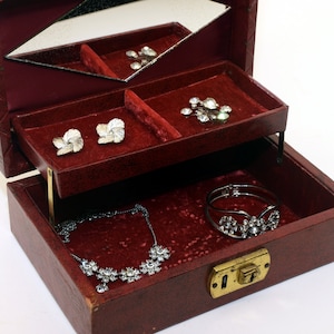Puede incluir: Una caja de joyería forrada de terciopelo rojo con una tapa con bisagras y un espejo en el interior. La caja tiene una cerradura y una llave y está llena de varias joyas, incluyendo un collar, una pulsera, pendientes y un broche.