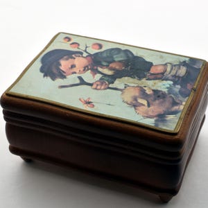 May include: A small, rectangular wooden box with a hinged lid. The lid features a colorful illustration of a child with a dog. The box is a dark brown color with a glossy finish and sits on four small feet. The illustration has a gold border.