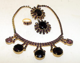 May include: A purple rhinestone necklace with a matching ring and two brooches. The necklace has five large round stones and a delicate chain. The brooches are both round with smaller rhinestones surrounding a larger centre stone.