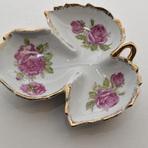 Puede incluir: Un plato de cerámica blanco en forma de hoja con tres secciones. Presenta diseños florales de rosas rosas y ribete dorado a lo largo de los bordes y el asa. El plato se utiliza probablemente para servir pequeñas porciones de comida o como objeto decorativo.