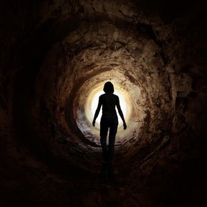 May include: A silhouette of a person walking towards a bright light at the end of a dark tunnel.