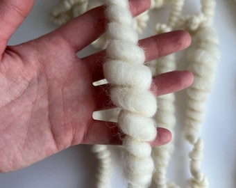 May include: A close-up of a white, fluffy yarn. The yarn is twisted into a rope-like shape and is held in a hand.
