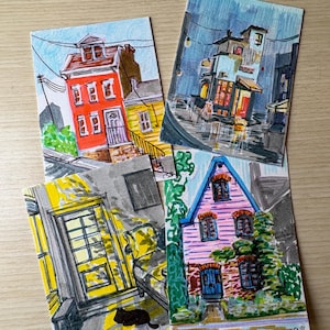 May include: Four rectangular art prints featuring hand-drawn illustrations of buildings. One shows a red house, another a bookstore at night, a third a yellow door with a black cat, and the fourth a pink house covered in green vines.