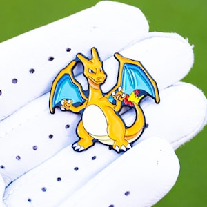 May include: A yellow and orange Charizard golf ball marker with blue wings and accents, held in a white golf glove. The marker is a detailed enamel design, set against a green background.