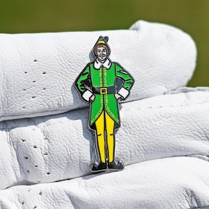 May include: A white leather golf glove with a Buddy the Elf enamel pin. The pin features Buddy in his green elf uniform with a yellow belt and yellow pants.