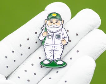May include: A colorful enamel pin featuring a cartoon gnome wearing a green hat and carrying a green bag. The gnome is wearing a white outfit with the number 10 on it. The pin is on a white golf glove against a green background.