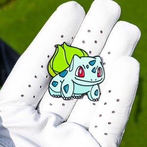 May include: A metal enamel pin featuring a light blue creature with a large green bulb on its back and red eyes. The character has dark blue spots on its body and is outlined in black, showcasing a cartoon design with a smooth, glossy finish.