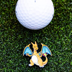 May include: A white golf ball with a dimpled surface rests above a colorful Charizard pin on a bed of green artificial turf. The pin features a yellow and orange dragon with blue wings, a popular character from the Pok&eacute;mon franchise.