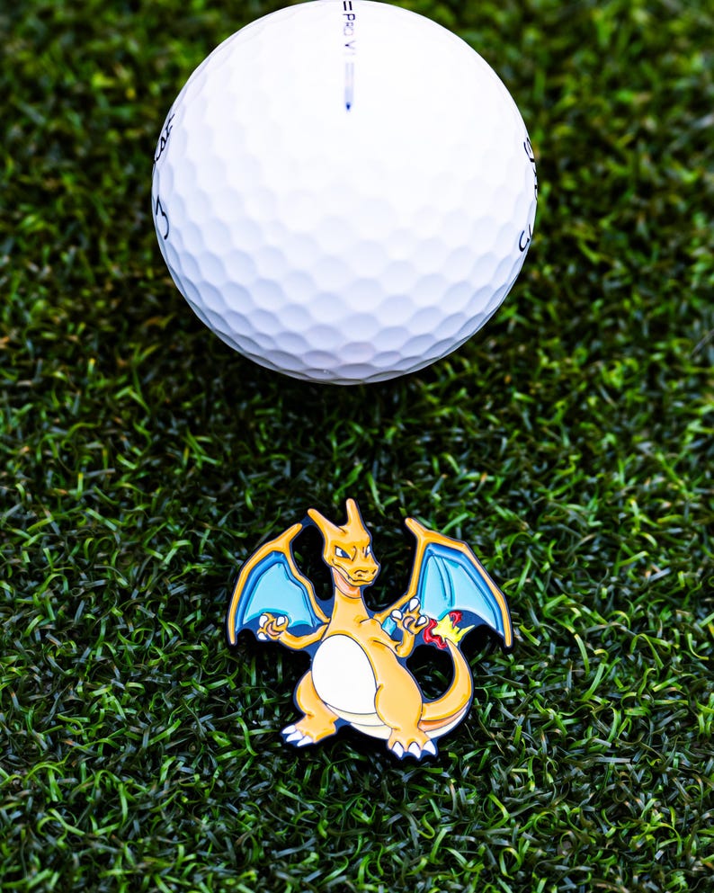 May include: A white golf ball with a dimpled surface rests above a colorful Charizard pin on a bed of green artificial turf. The pin features a yellow and orange dragon with blue wings, a popular character from the Pok&eacute;mon franchise.