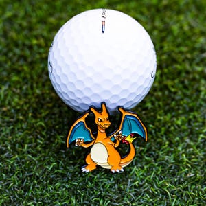 May include: A white golf ball with a Charizard-themed ball marker. The marker features an orange and yellow Charizard design with blue wings. The golf ball and marker are set on a green surface.