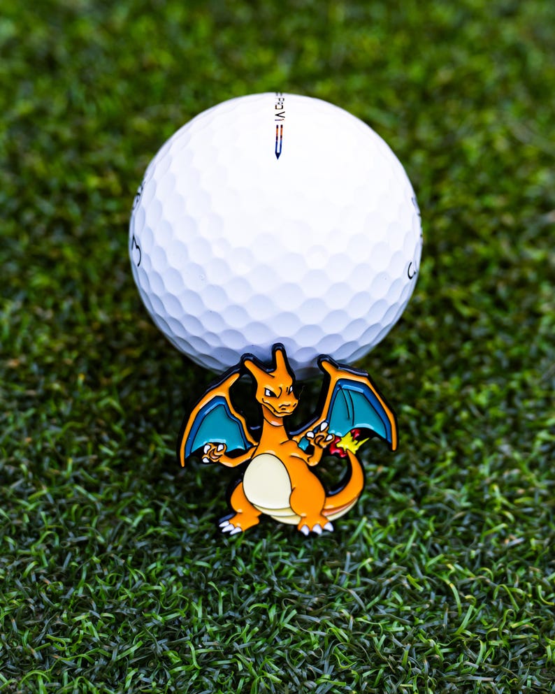 May include: A white golf ball with a Charizard-themed ball marker. The marker features an orange and yellow Charizard design with blue wings. The golf ball and marker are set on a green surface.