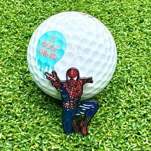 K&ouml;nnte beinhalten: Ein wei&szlig;er Golfball mit einem blauen und wei&szlig;en Aufkleber, auf dem "A Golf Thing" steht, und einer roten und blauen Spiderman-Anstecknadel.