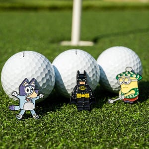 Puede incluir: Tres pelotas de golf blancas con hoyuelos dispuestas en fila, cada una con un pin de esmalte de colores en la parte delantera. Los pines representan a un perro azul de dibujos animados, un superhéroe negro y amarillo y un minion con sombrero verde.