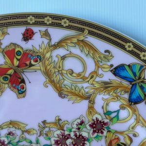 May include: A decorative plate with a butterfly and floral design. The plate features a gold border with a repeating geometric pattern. Butterflies in red, blue, and yellow are depicted among gold-colored foliage and flowers.
