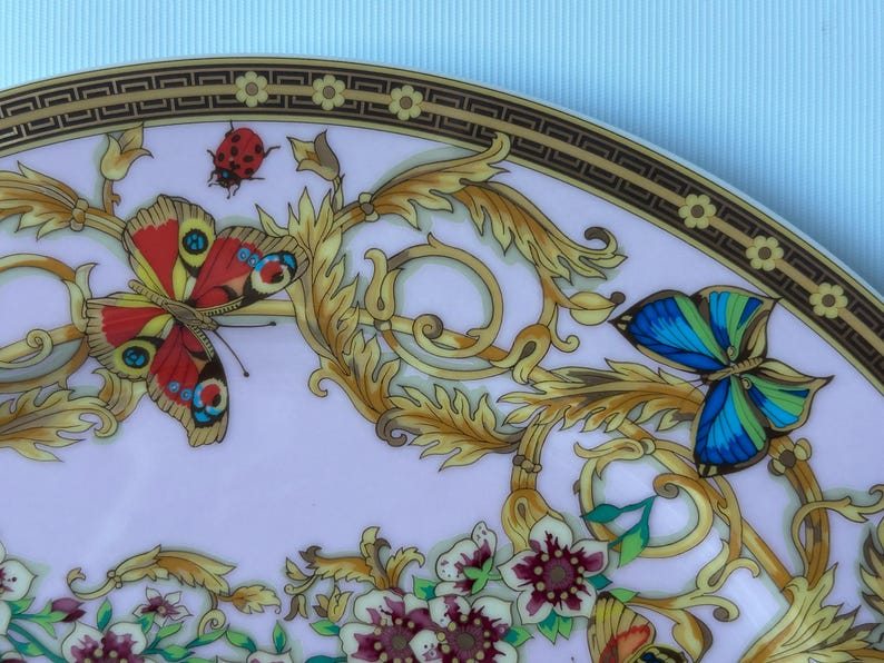 May include: A decorative plate with a butterfly and floral design. The plate features a gold border with a repeating geometric pattern. Butterflies in red, blue, and yellow are depicted among gold-colored foliage and flowers.
