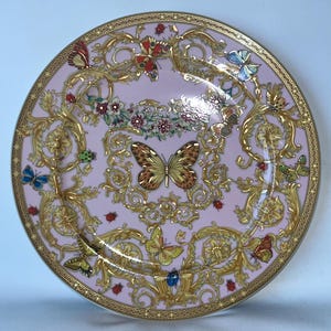 May include: A decorative pink plate with a gold rim and ornate gold detailing. The plate features butterflies, ladybugs, and floral accents. The design is symmetrical and elegant, suitable for display or dining.