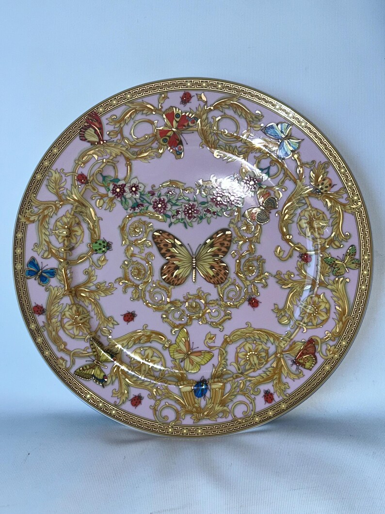 May include: A decorative pink plate with a gold rim and ornate gold detailing. The plate features butterflies, ladybugs, and floral accents. The design is symmetrical and elegant, suitable for display or dining.