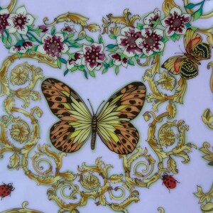May include: A decorative plate with a light purple background, featuring a butterfly with brown and yellow wings, surrounded by gold scrollwork, flowers, and ladybugs. The plate has a floral and insect theme.