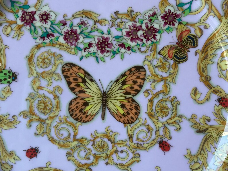 May include: A decorative plate with a light purple background, featuring a butterfly with brown and yellow wings, surrounded by gold scrollwork, flowers, and ladybugs. The plate has a floral and insect theme.