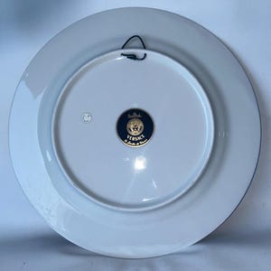 May include: A white ceramic plate with a blue border and a gold and navy blue Versace emblem. The plate has a black wire for hanging. The back of the plate is visible.