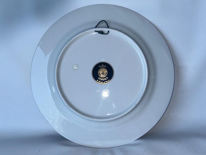 May include: A white ceramic plate with a blue border and a gold and navy blue Versace emblem. The plate has a black wire for hanging. The back of the plate is visible.