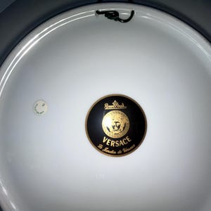May include: White, oval-shaped decorative plate with a black and gold Versace emblem in the center. The emblem features the Versace logo and the text "Le Jardin de Versace". A small, circular sticker is visible on the left side.
