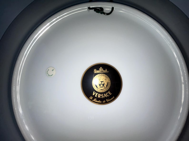 May include: White, oval-shaped decorative plate with a black and gold Versace emblem in the center. The emblem features the Versace logo and the text "Le Jardin de Versace". A small, circular sticker is visible on the left side.