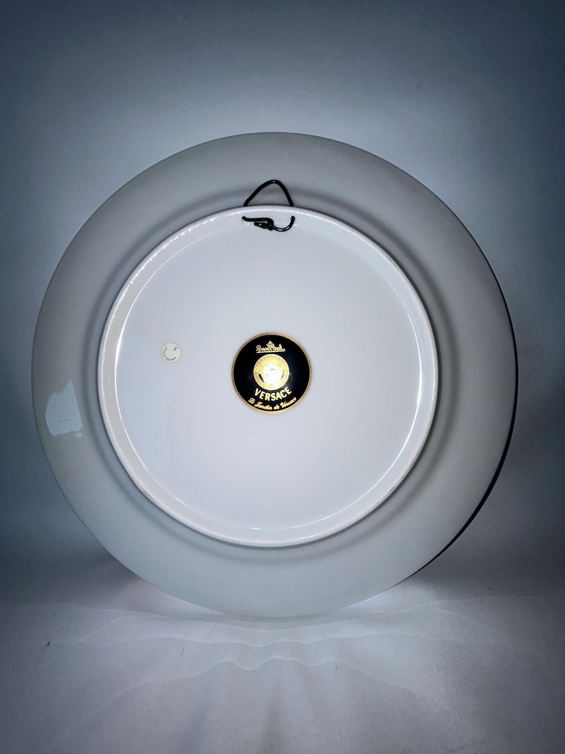 May include: Back of a white porcelain decorative plate with a gray rim. A black metal hanger is attached to the top. The center features a black and gold Versace logo.