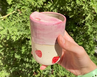 Könnte beinhalten: Eine rosa-weiße Keramiktasse mit Erdbeer-Design. Die Tasse hat einen Deckel mit einer kleinen Öffnung zum Trinken.