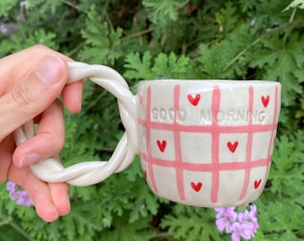 May include: A ceramic mug with a twisted handle. The mug is off-white with a pink grid pattern and small red hearts. The words "GOOD MORNING" are printed on the side. The mug is held against a backdrop of green foliage.