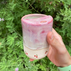 Puede incluir: Una taza de cerámica rosa y blanca con un diseño de cerezas. La taza tiene un pequeño agujero en la parte superior para una pajita.